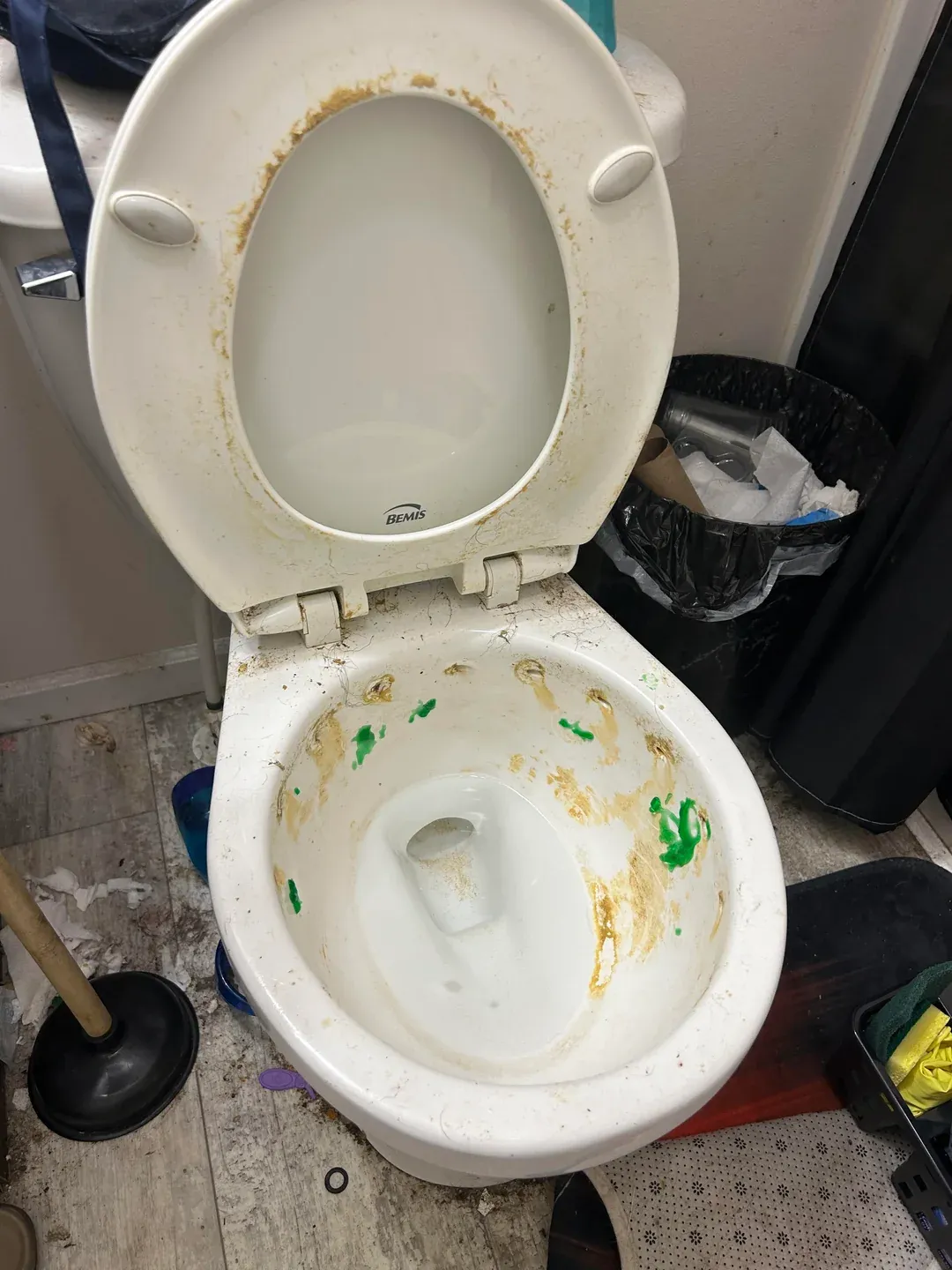 Dirty toilet bowl with brown and green stains, open lid, and surrounding clutter including a plunger and trash can in Drexel Heights, AZ