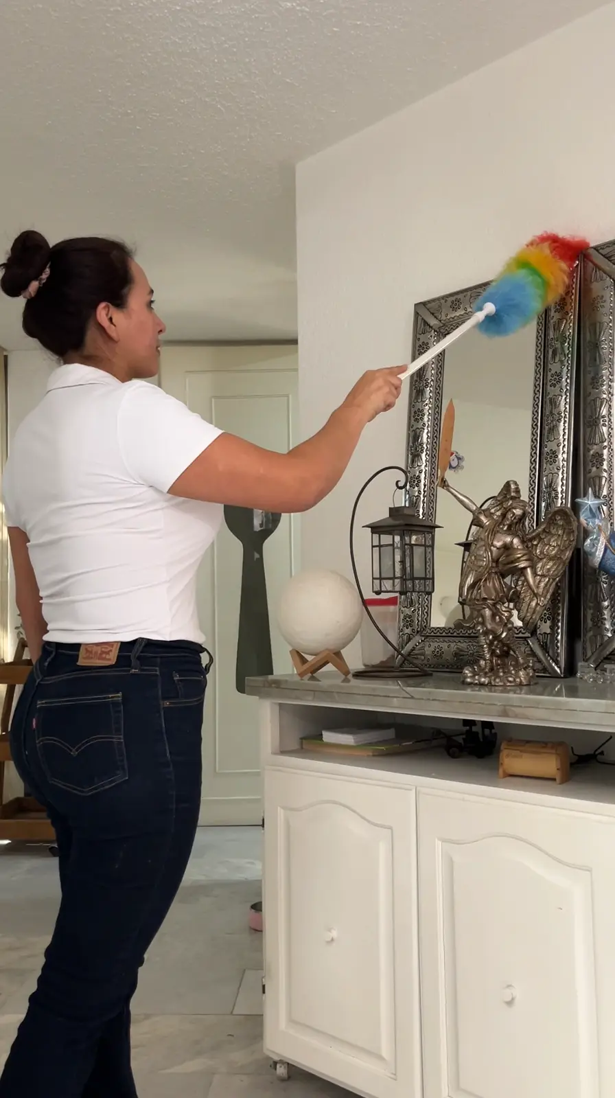 Woman dusting a silver-framed mirror with a rainbow-colored duster in a room with white furniture.
