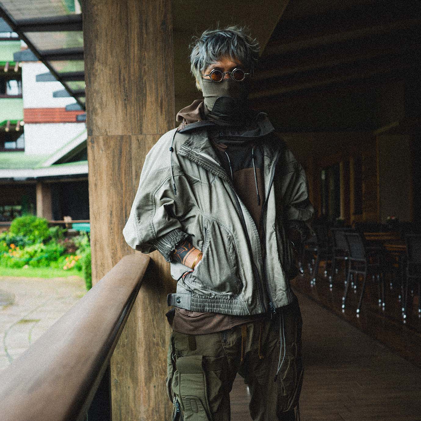 Person with gray hair wearing round glasses, a face mask, an oversized light jacket, and cargo pants standing indoors near a wooden railing.