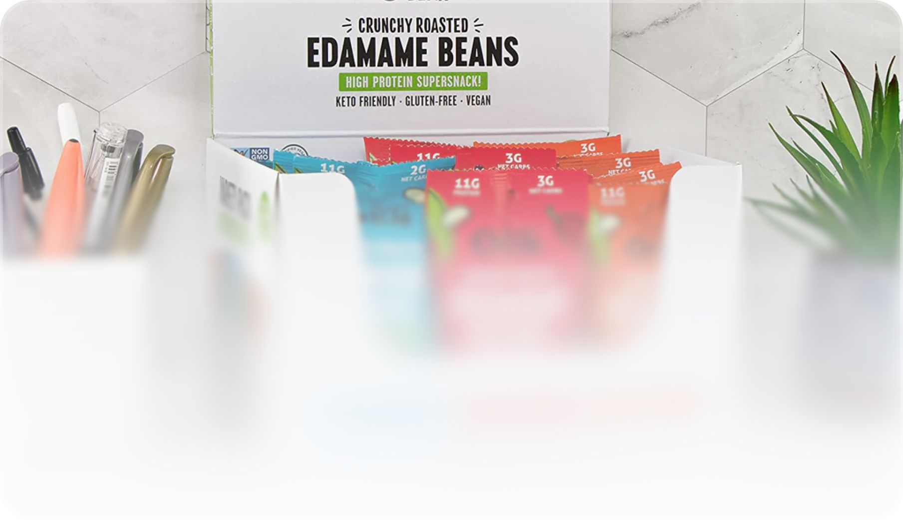 Display box of crunchy roasted edamame beans snack packets in various colors on a marble countertop with a pen holder and a small potted plant nearby.