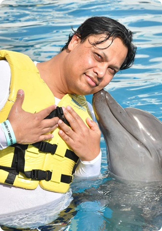 Man in a yellow life jacket smiling as a dolphin touches his cheek in a pool.