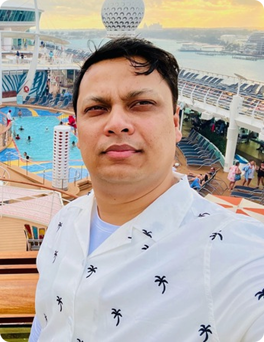 Man wearing a white shirt with black palm tree prints taking a selfie on a cruise ship deck with a pool and lounge chairs behind him at sunset.