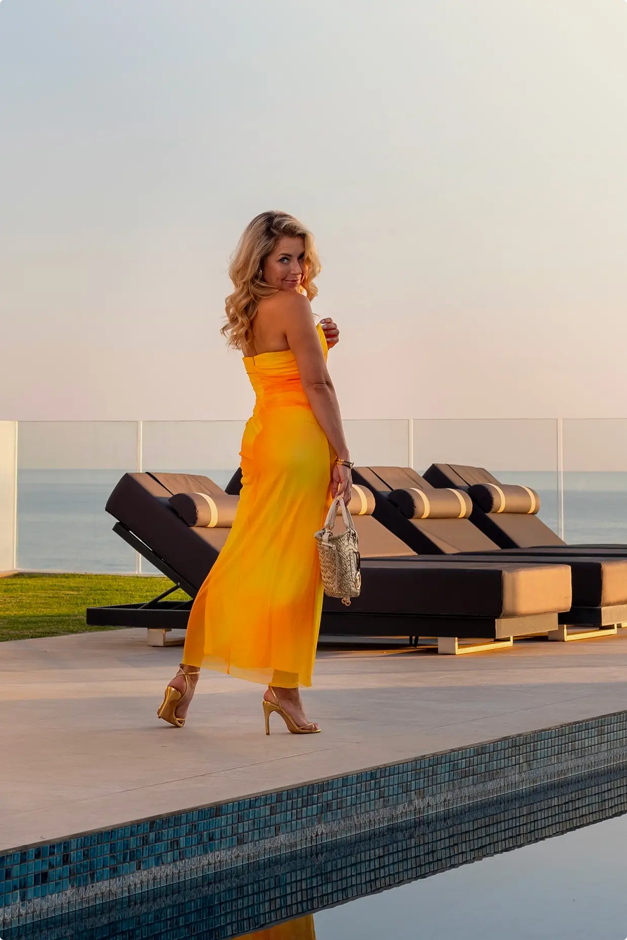Woman in a strapless yellow dress and gold heels holding a handbag walking by a pool with lounge chairs and an ocean view at sunset.
