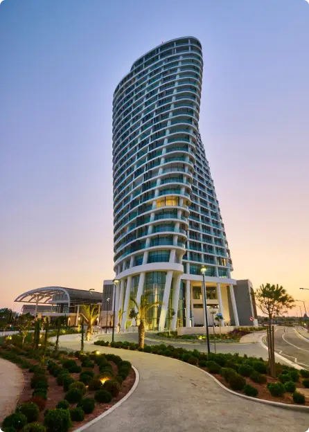 Tall modern curved glass building at dusk with landscaped pathway and palm trees in front.