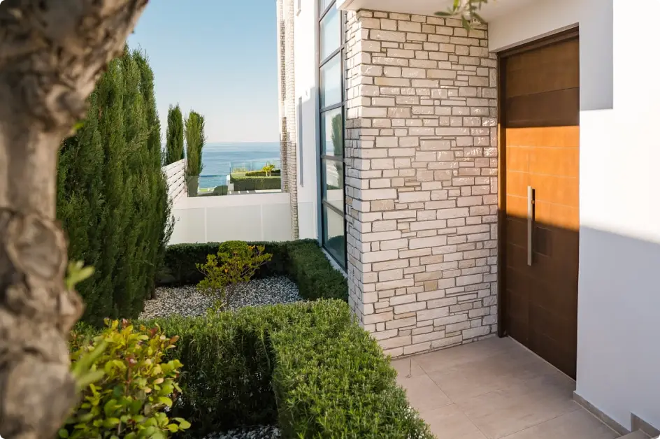 Modern house entrance with a wooden door, tiled floor, manicured green bushes, and a view of the ocean in the background.
