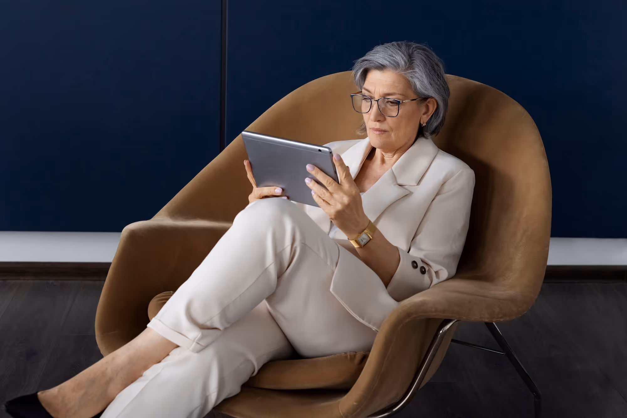 Older woman with gray hair and glasses sitting in a brown chair, reading on a tablet.