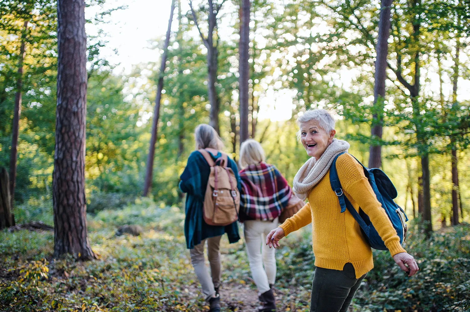 Leende äldre kvinna i gul tröja och halsduk med ryggsäck, promenerande i en skog tillsammans med två andra kvinnor.