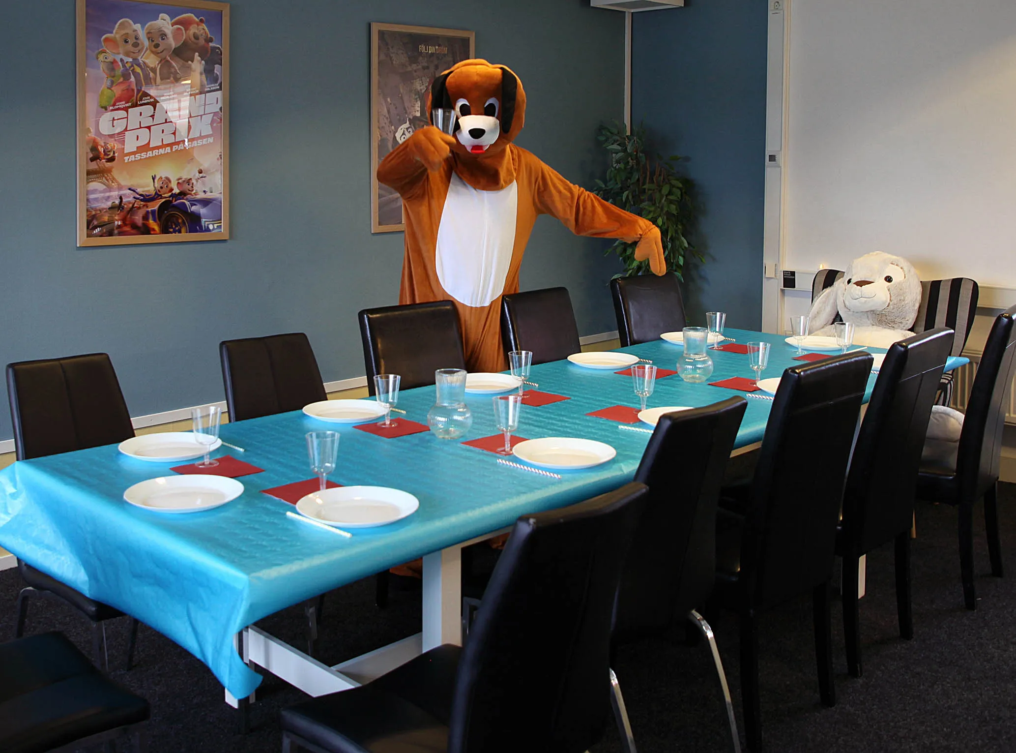 Ett barnkalasbord med blå duk, vita tallrikar, glas och röda servetter, med en person i hundmaskotdräkt och en stor mjukisdjurskanin i rummet på på barnkalas på KalasHuset i Nyköping.