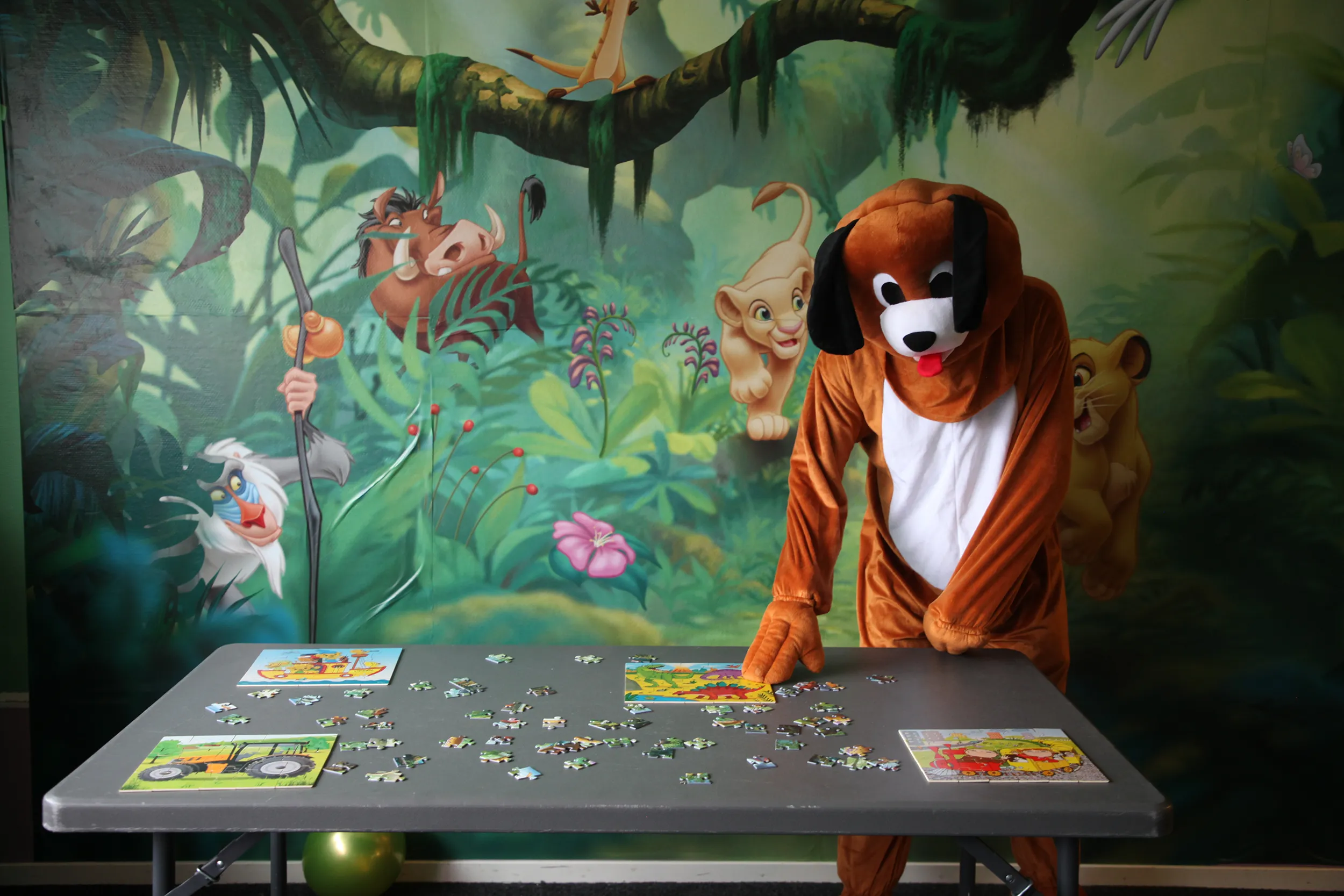 Person i hunddräkt står vid ett bord med pusselbitar framför en väggmålning med tecknade djurfigurer från Lejonkungen. Pussel på barnkalas på KalasHuset i Nyköping.