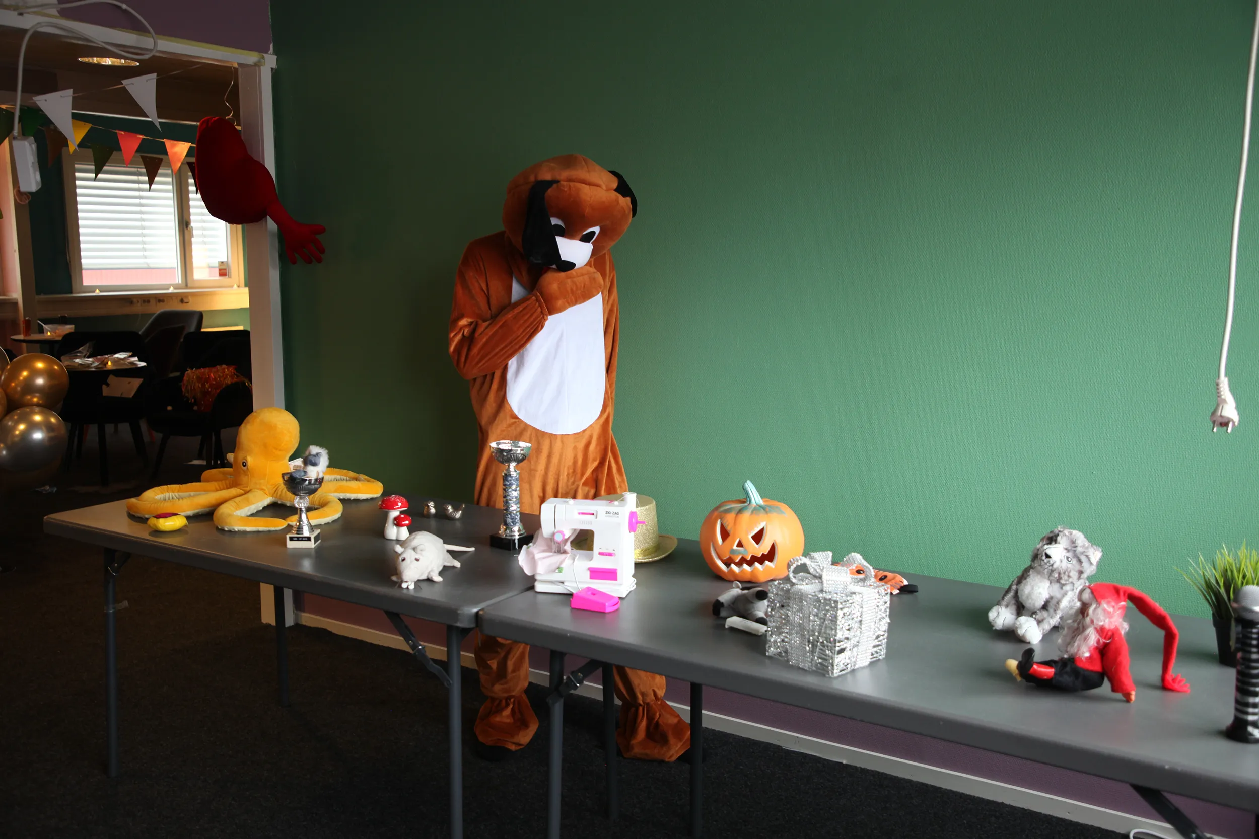 Person i hunddräkt står vid ett bord med halloween- och leksaksföremål inklusive pumpa, gosedjur och pris på barnkalas på KalasHuset i Nyköping.