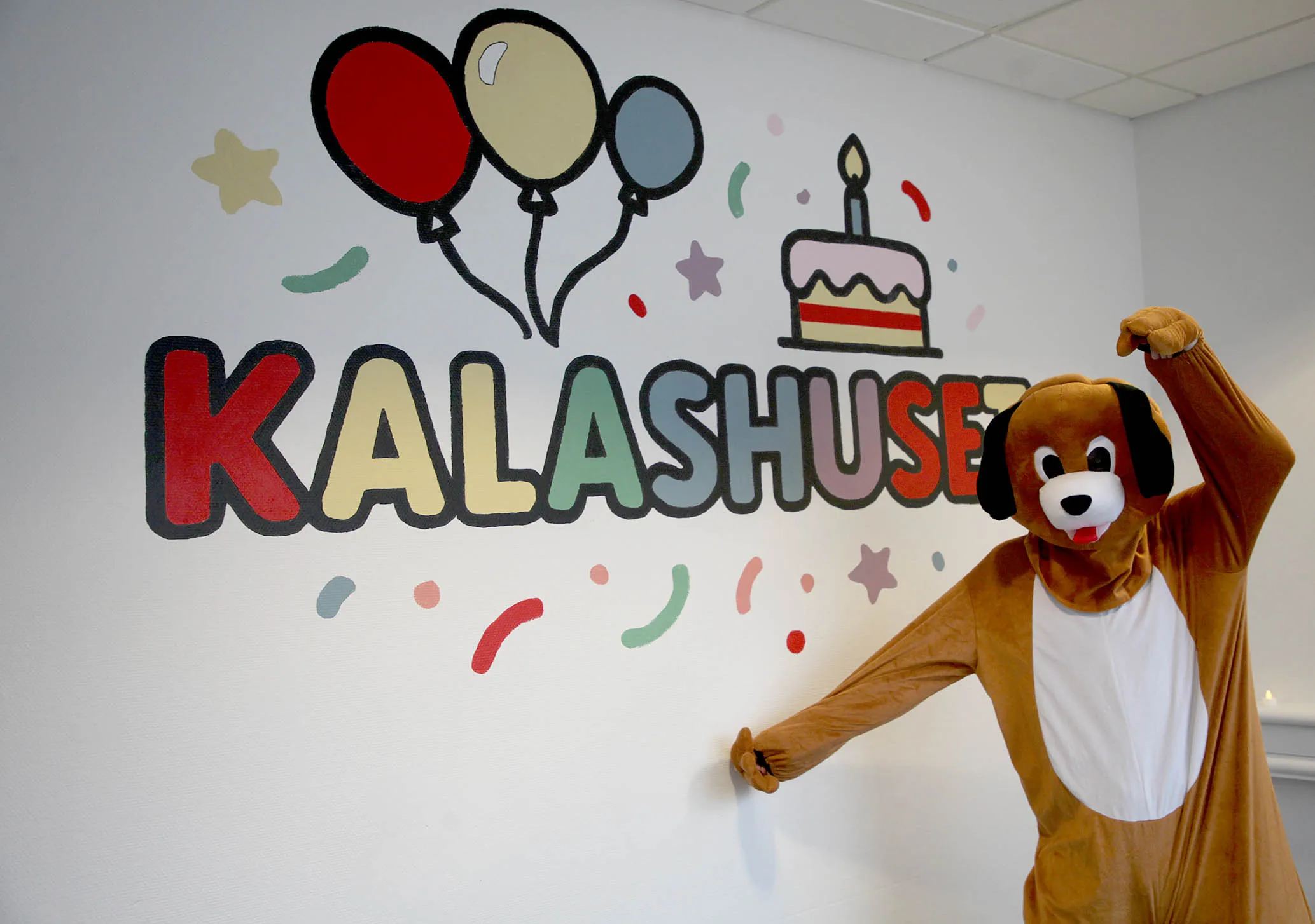 Person i hunddräkt poserar bredvid en väggmålning med texten Kalashuset, ballonger och en tårta med ett ljus på barnkalas på KalasHuset i Nyköping.