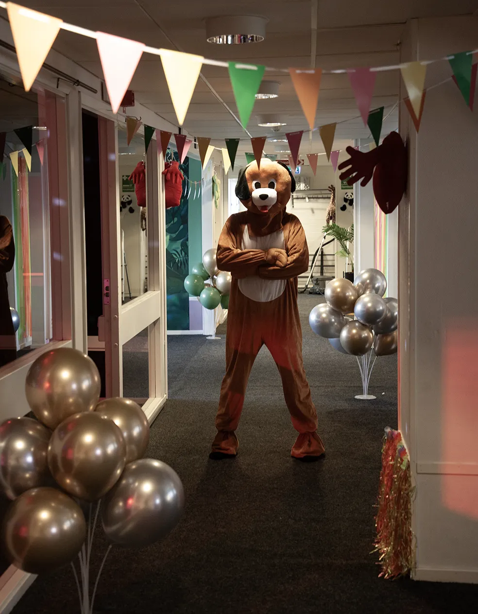 Person i hunddräkt står i en festdekorerad korridor med färgglada vimplar och glimmande ballonger på barnkalas på KalasHuset i Nyköping.