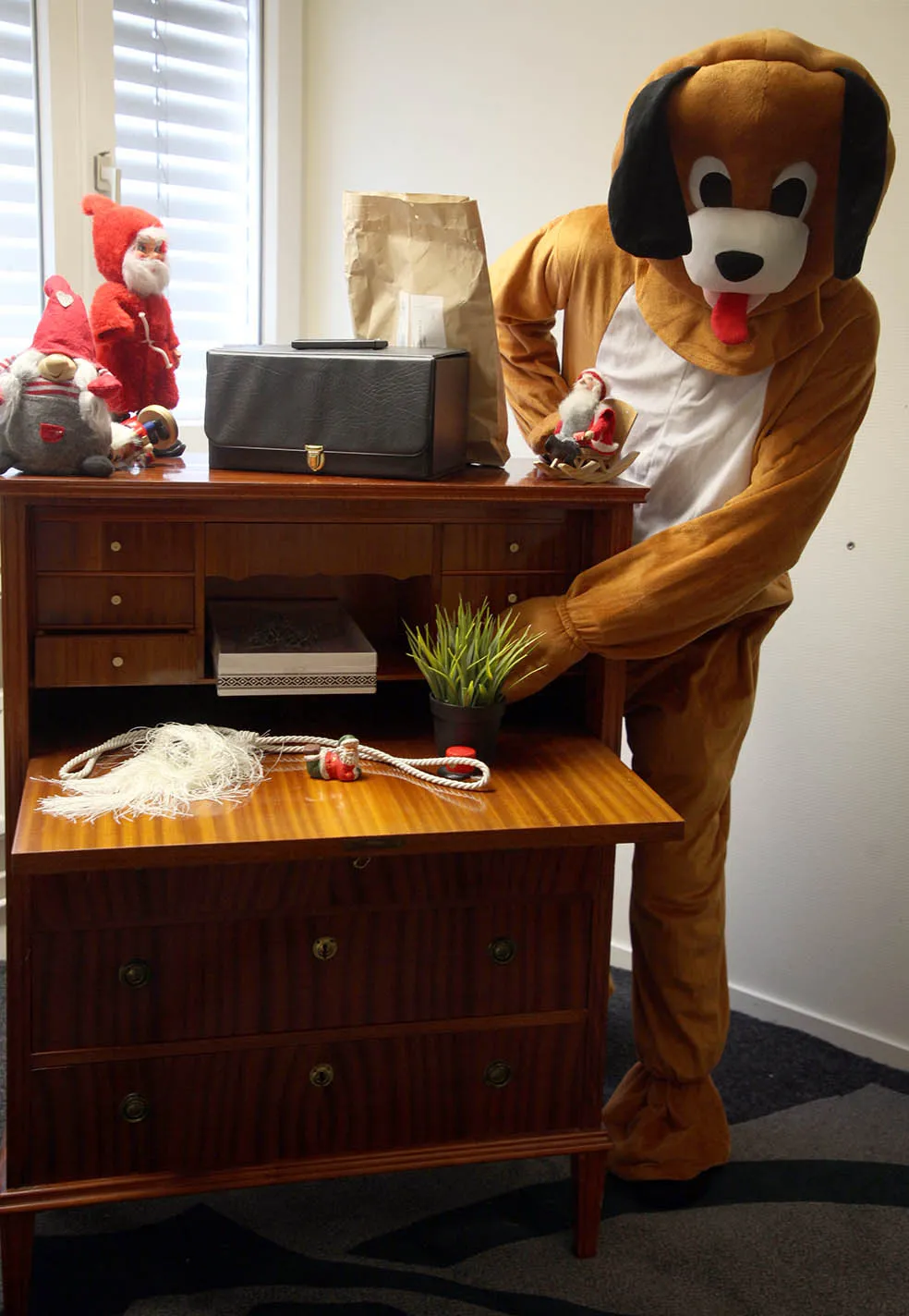 Person i hunddräkt som ställer en krukväxt på en öppen träbyrå med tomtar och andra föremål ovanpå på barnkalas på KalasHuset i Nyköping.