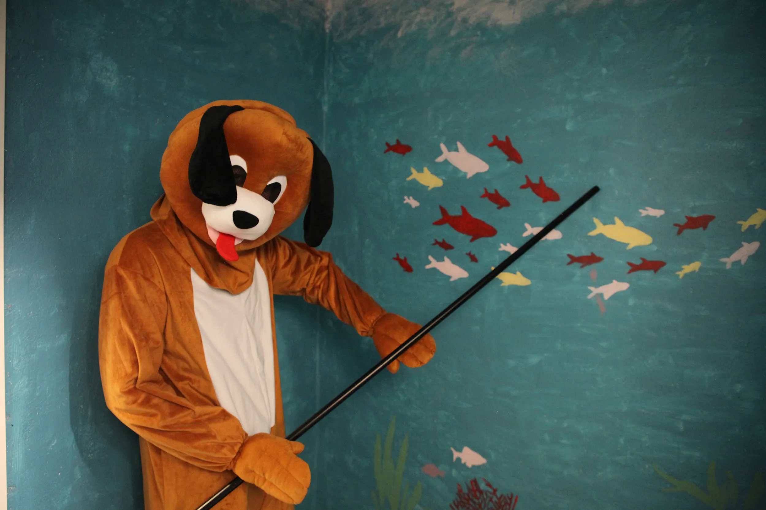 Person i hunddräkt pekar med en lång pinne mot målade fiskar på en blå vägg. Fiskdamm på barnkalas på KalasHuset i Nyköping.