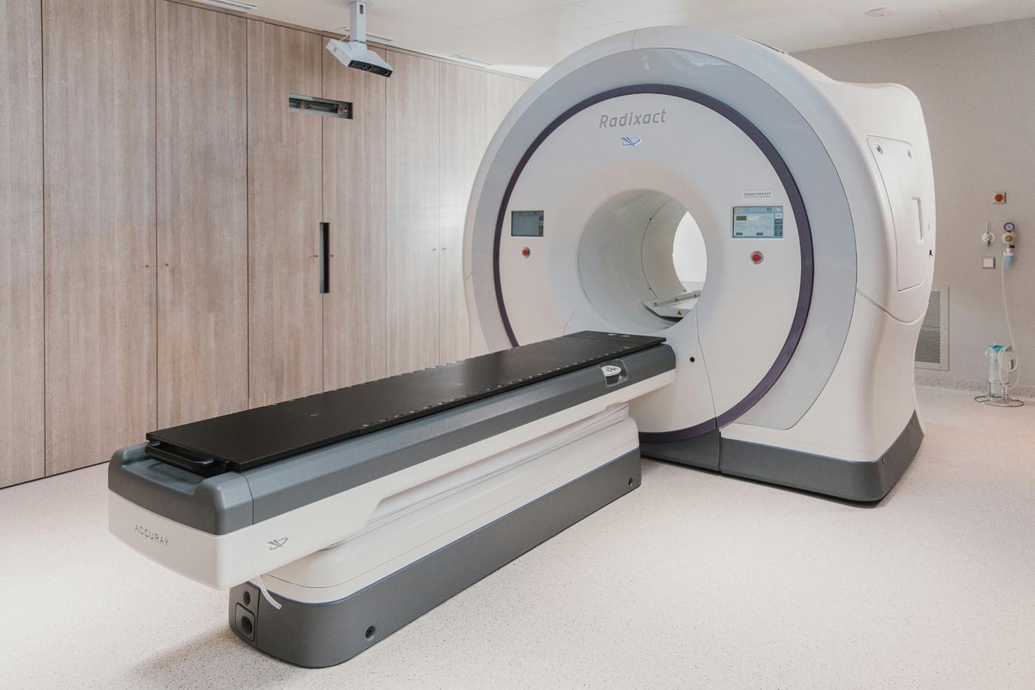 Modern medical radiotherapy machine in a clean treatment room with wooden cabinets in the background.