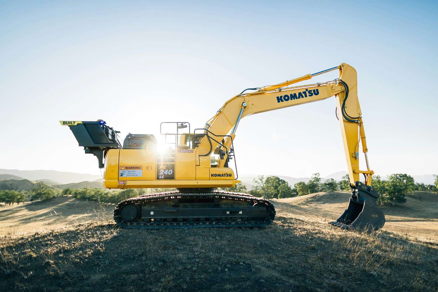 Yellow Komatsu excavator with a black digging bucket on dry grassland with hills and trees in the background.