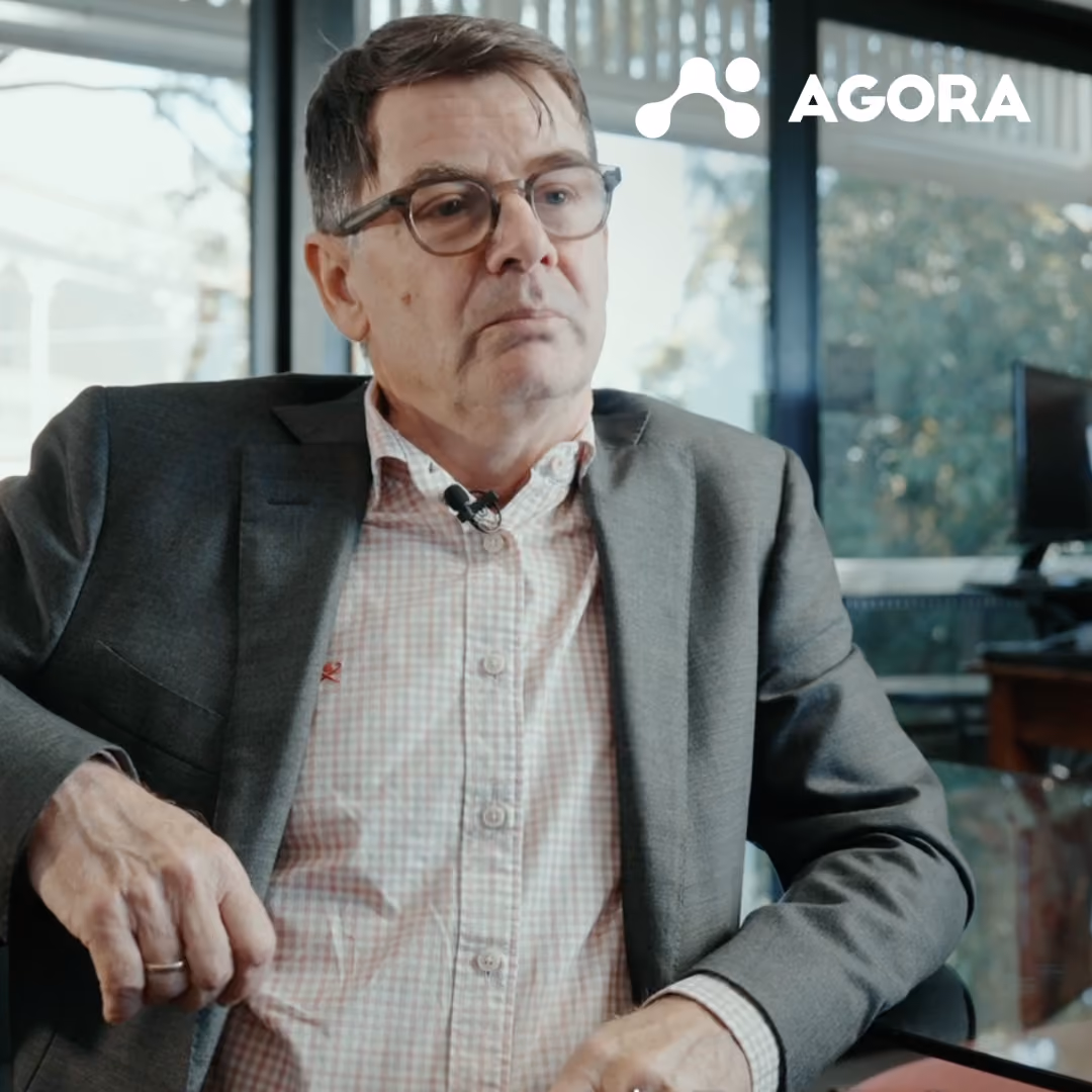 Man in glasses wearing a gray suit and checked shirt sitting in an office with large windows.