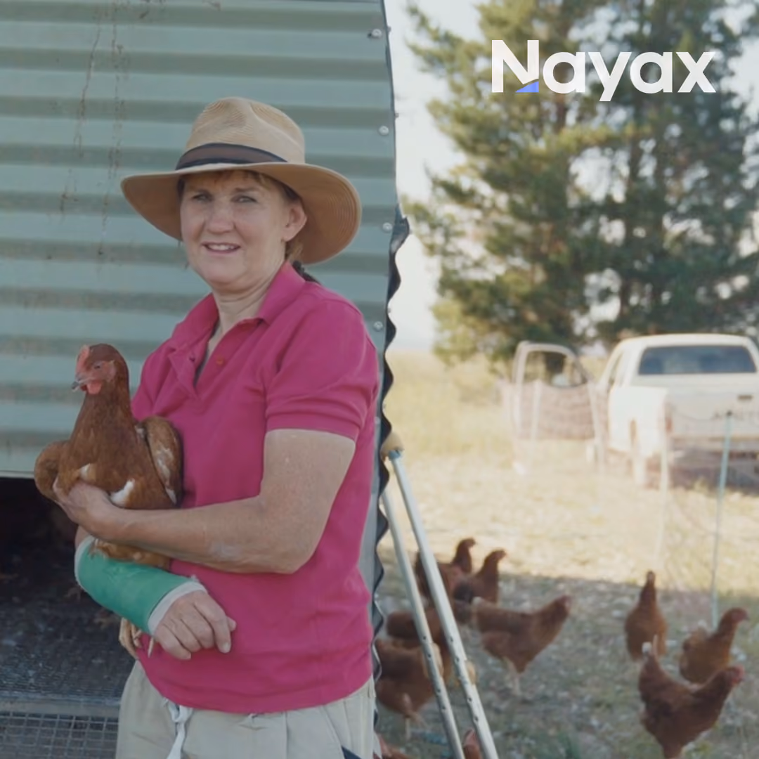Woman in a pink shirt and hat holding a brown chicken with crutches and chickens outdoors behind her.