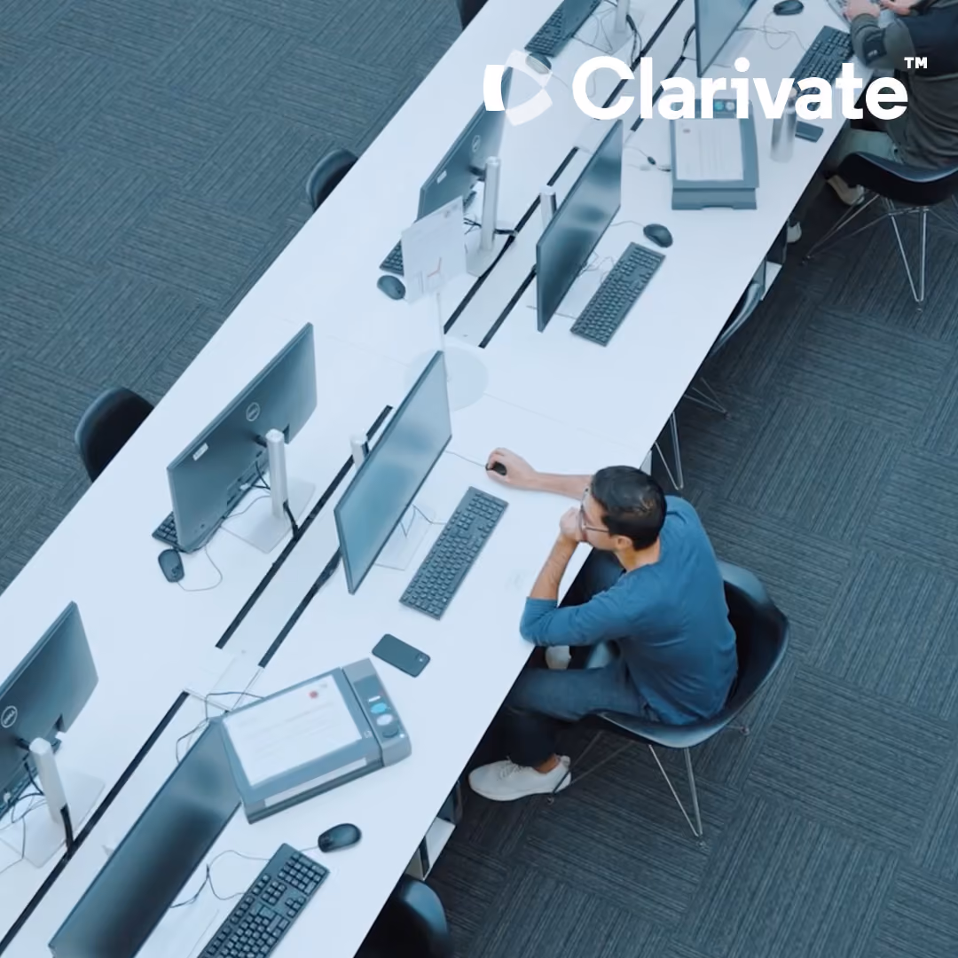 Overhead view of two people working at computers in a modern office with Clarivate logo.