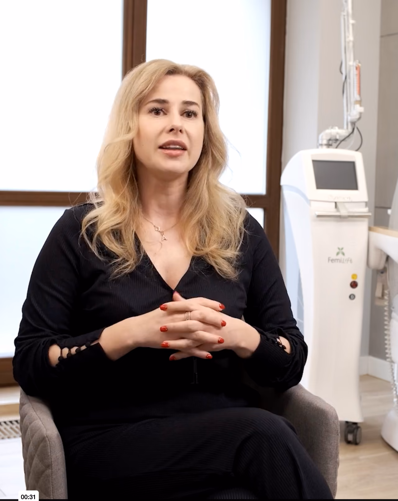 Blonde woman with red nails sitting in an office chair wearing a black outfit, medical device in the background.