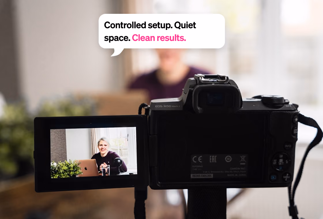 Camera screen shows a person sitting at a desk with a laptop and microphone in a bright, quiet room.