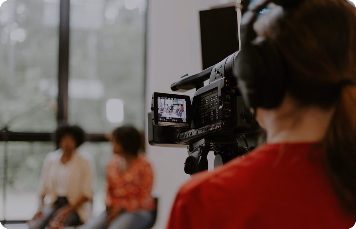 Camera operator filming two people seated and talking in front of large windows during a video shoot.