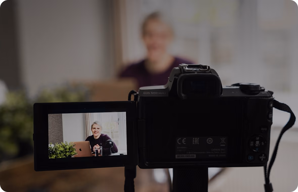 Viewfinder of a camera showing a woman smiling while sitting at a desk with a laptop and microphone.