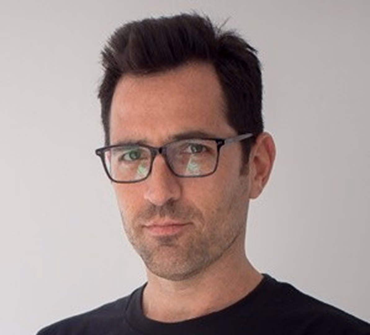 Man with dark hair, glasses, and stubble, wearing a black shirt against a plain light background.