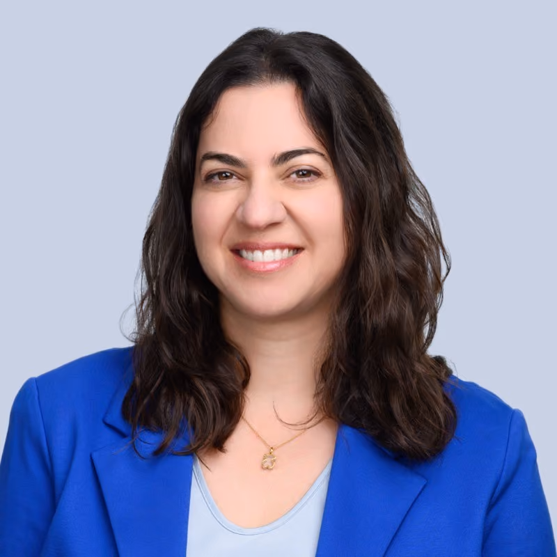 Smiling woman with shoulder-length dark hair wearing a blue blazer and light blue top against a gray background.