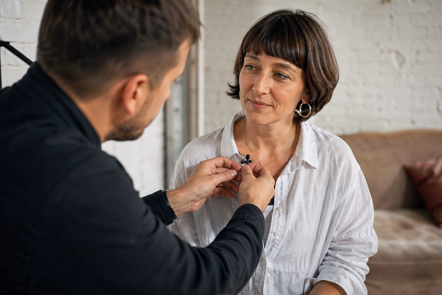 Man attaching a small microphone to a woman’s white shirt collar in a cozy room.