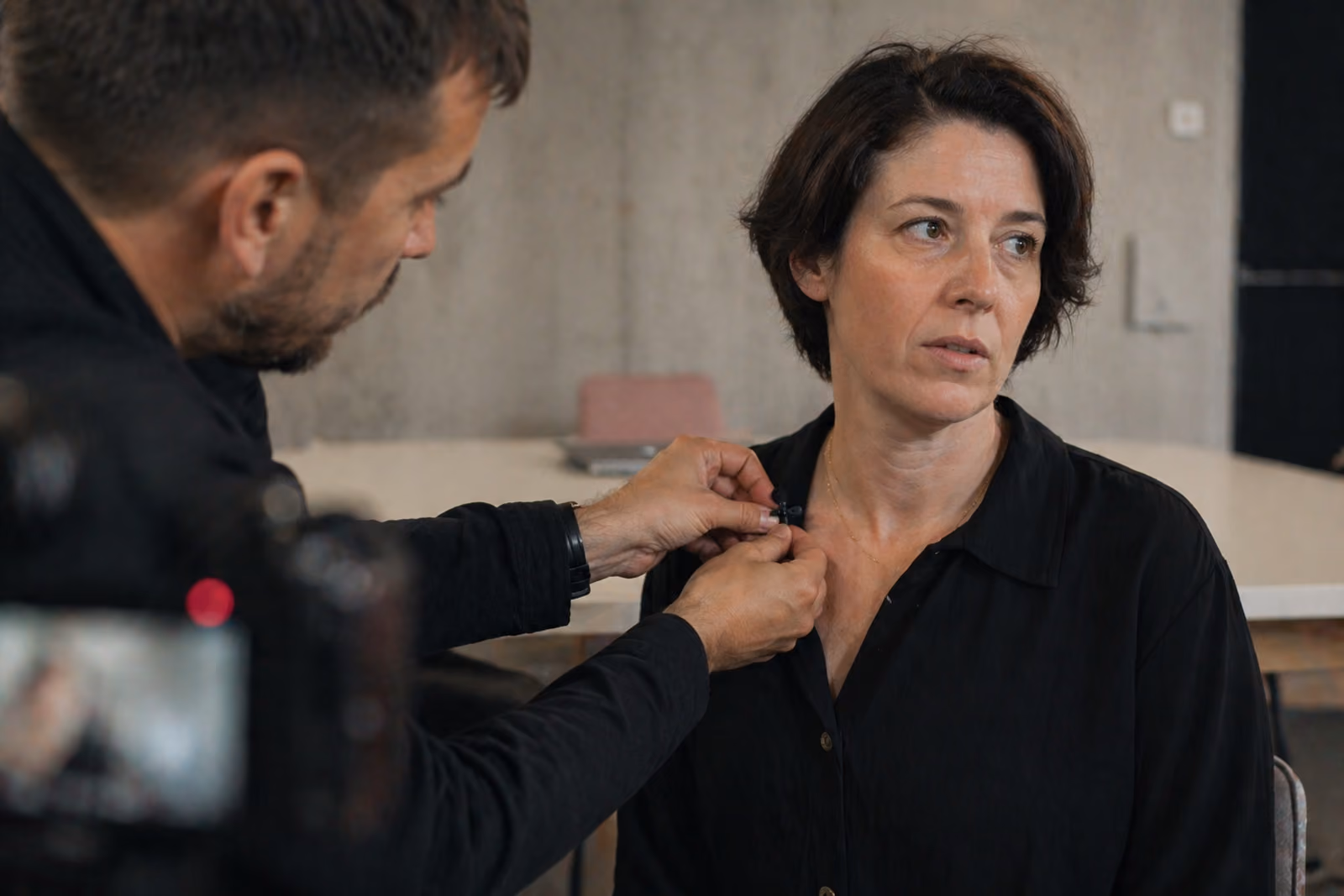 Man attaches a microphone to a woman’s black buttoned shirt in an indoor setting.