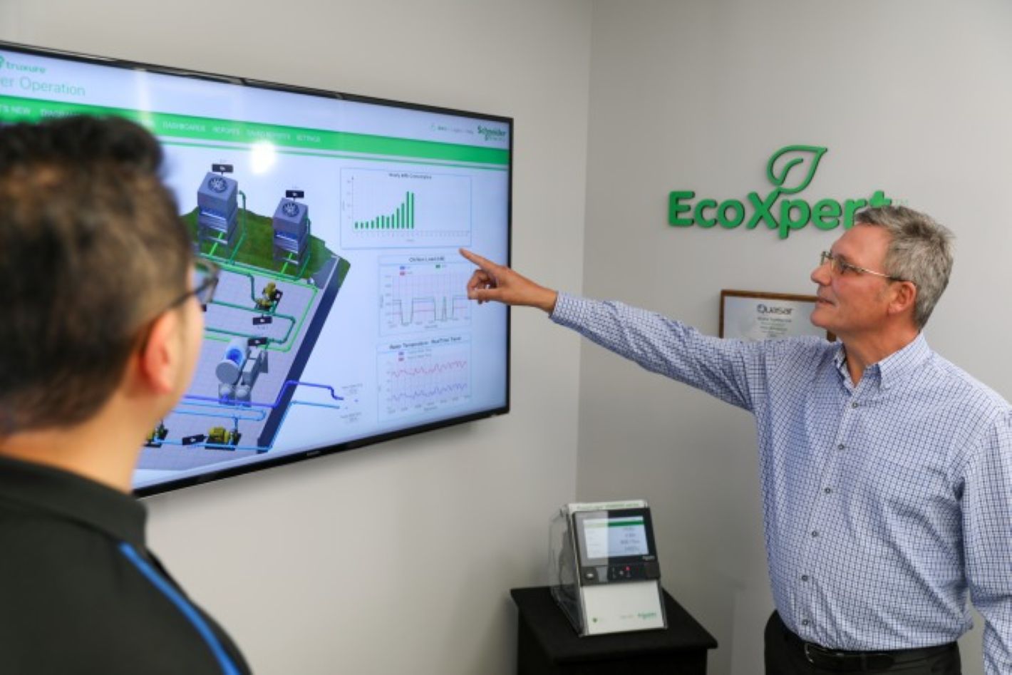 Man pointing at a screen displaying charts and diagrams while another person watches in a room with EcoXpert logo.