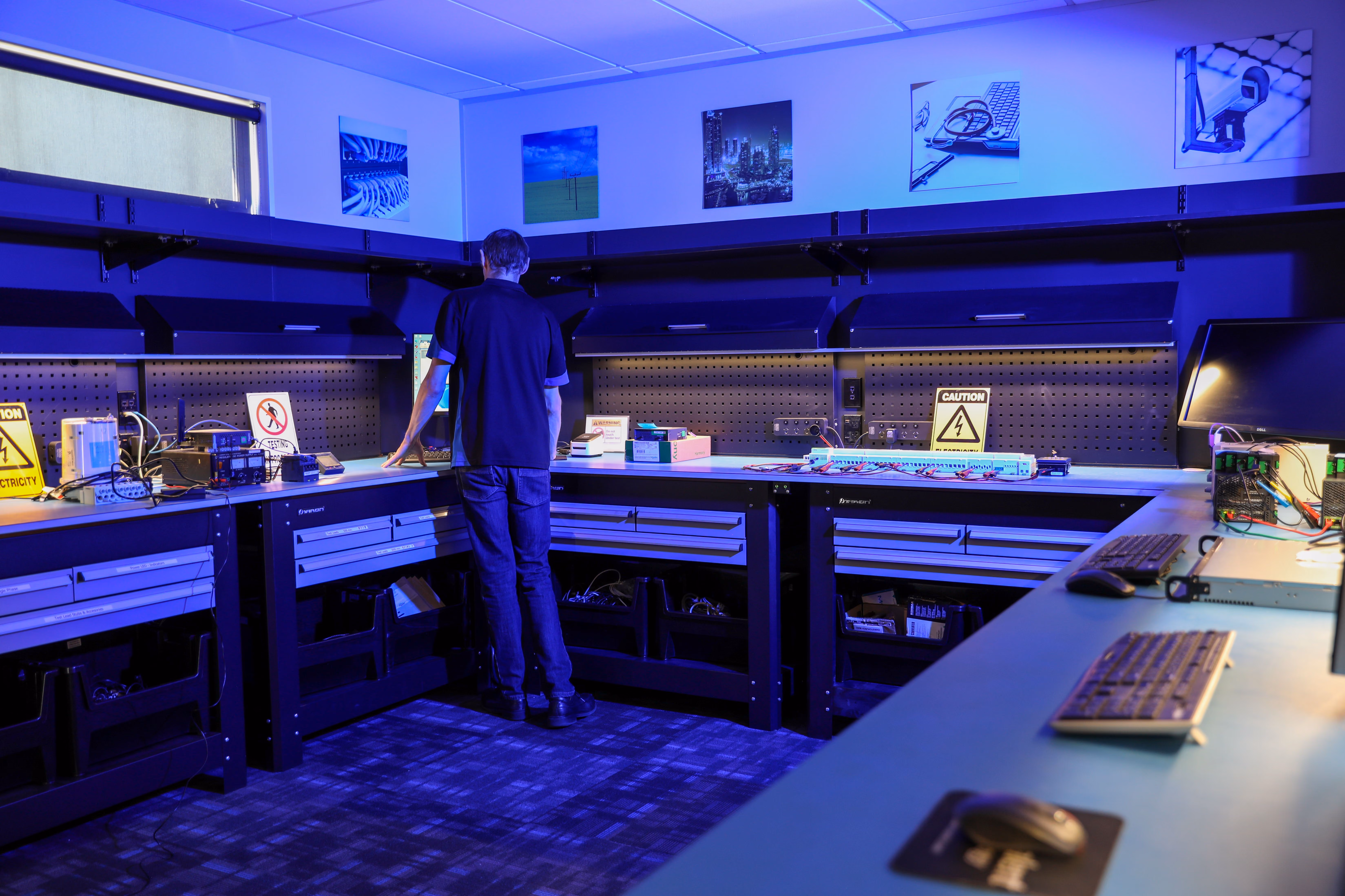 Man working at a computer workstation in a blue-lit electronics lab with caution signs and equipment.