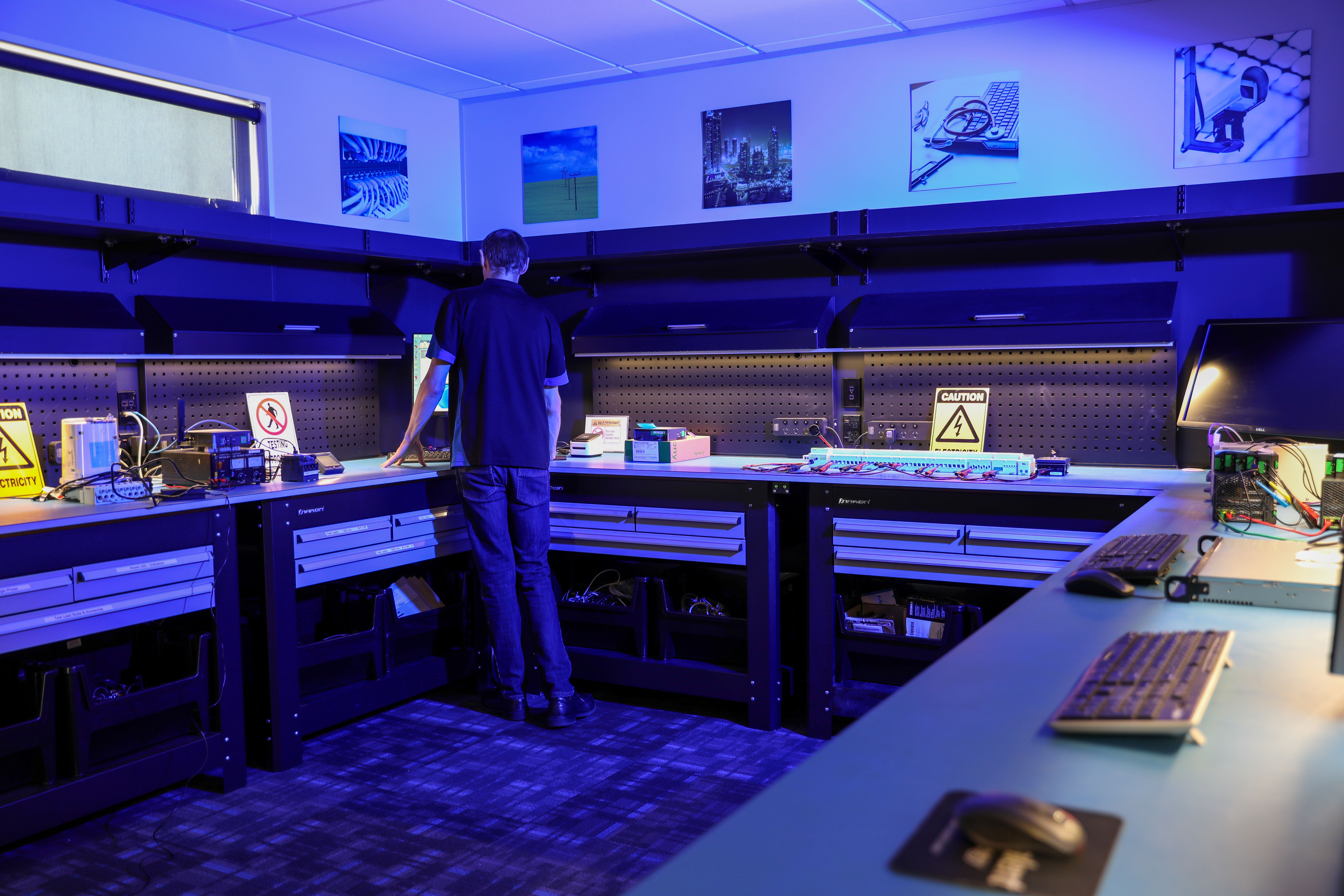 Man working at a computer workstation in a blue-lit electronics lab with caution signs and equipment.