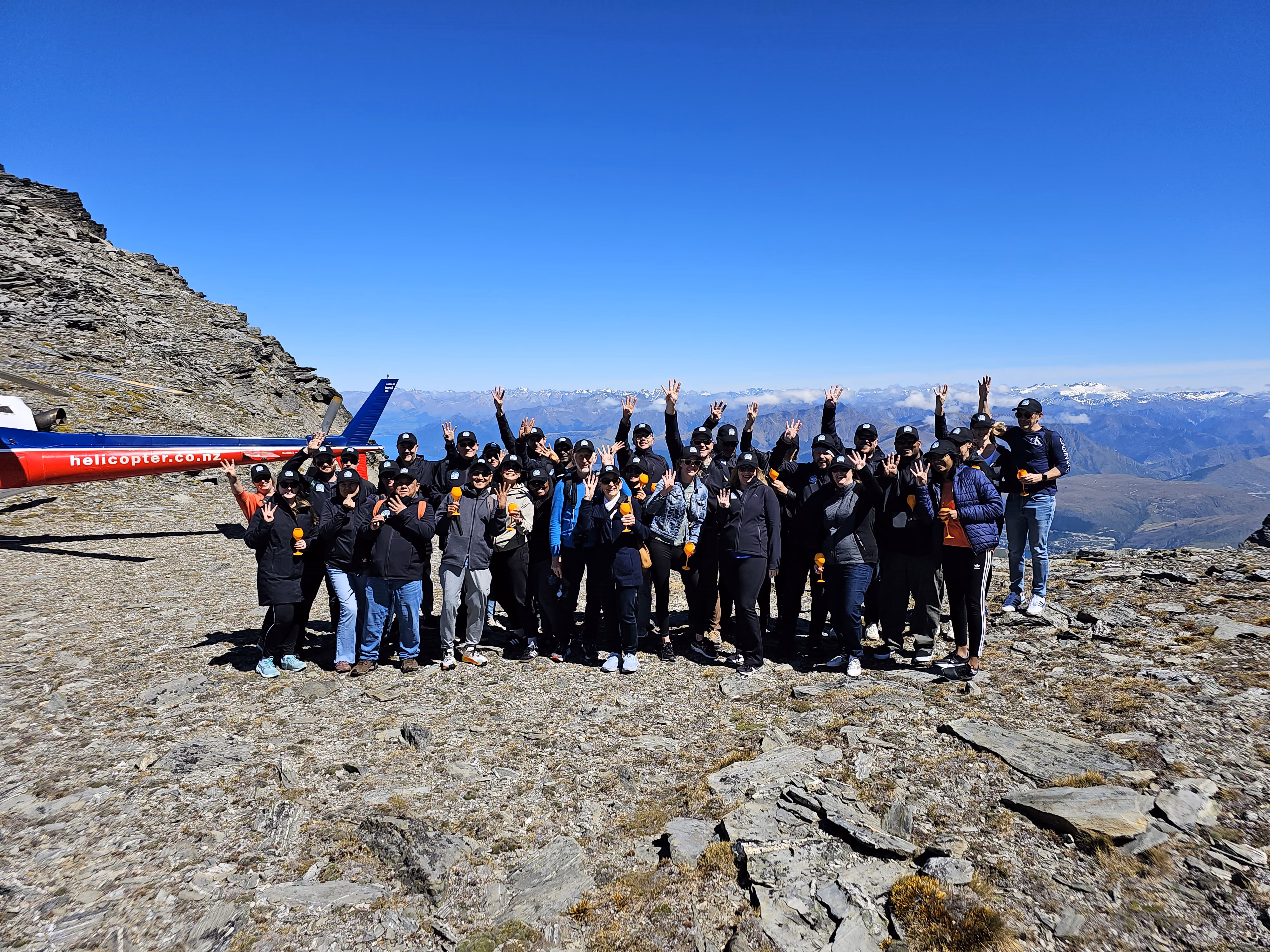 Group of people on rocky mountain with a helicopter and snowy peaks in background, many holding orange drinks.