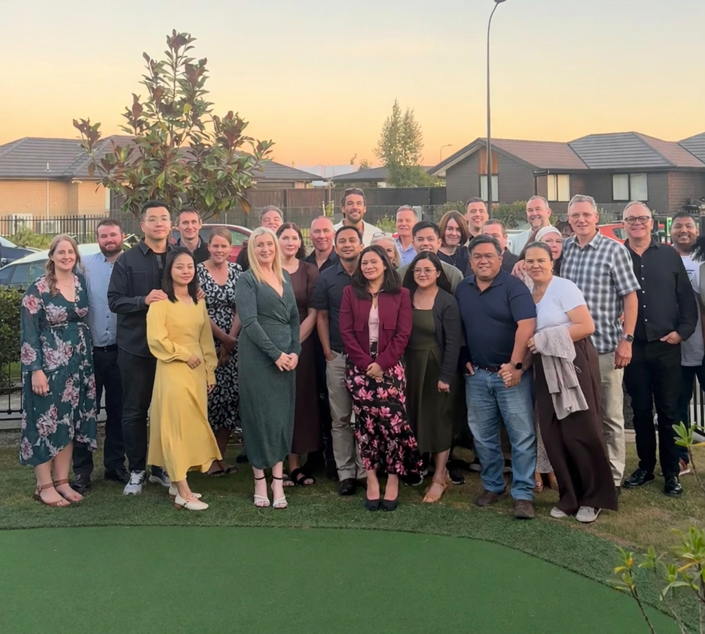 Large group of diverse adults posing outdoors on grass with houses and sunset in background.