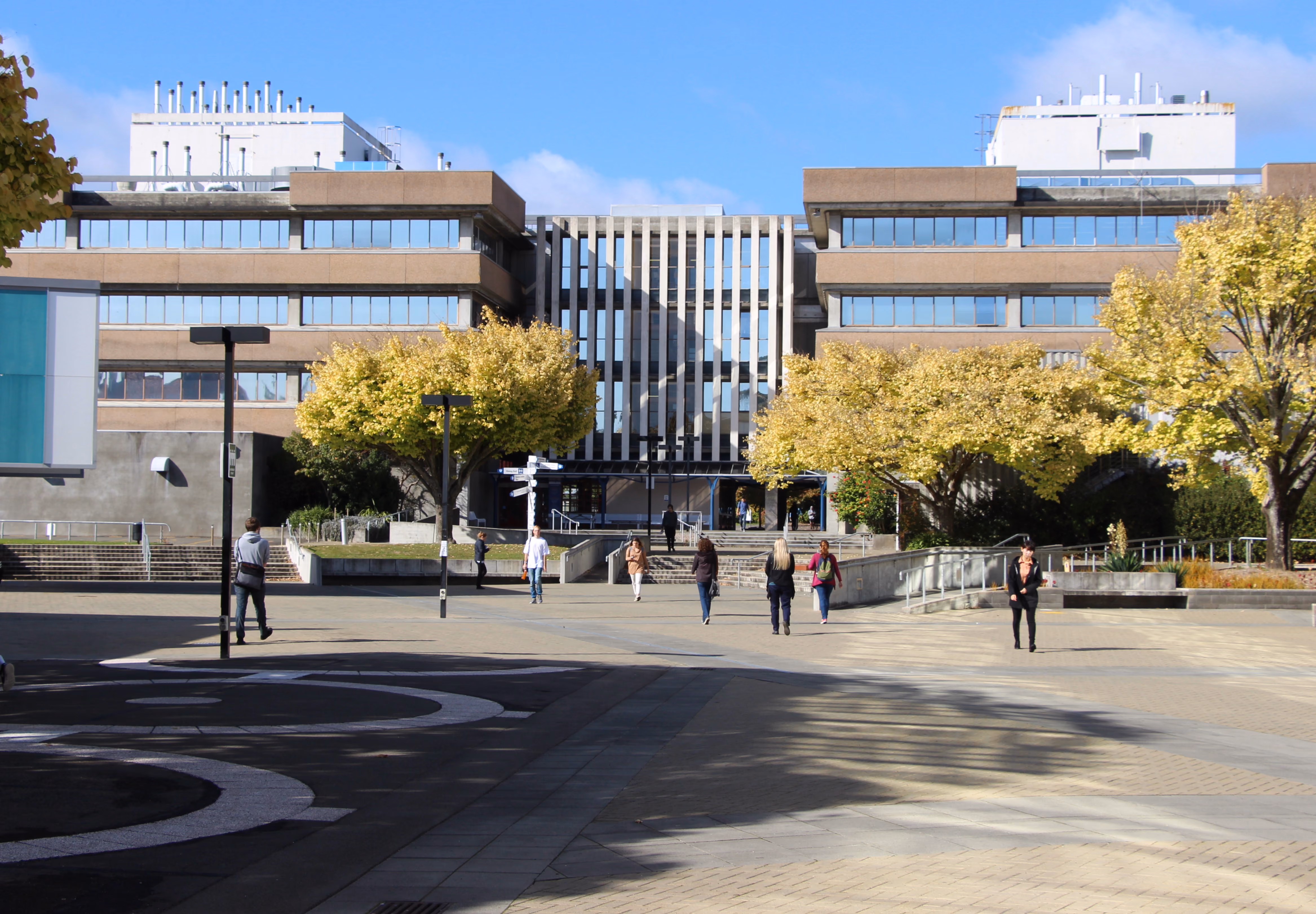 Wide concrete plaza with people walking toward a large modern building with yellow-leaved trees nearby.