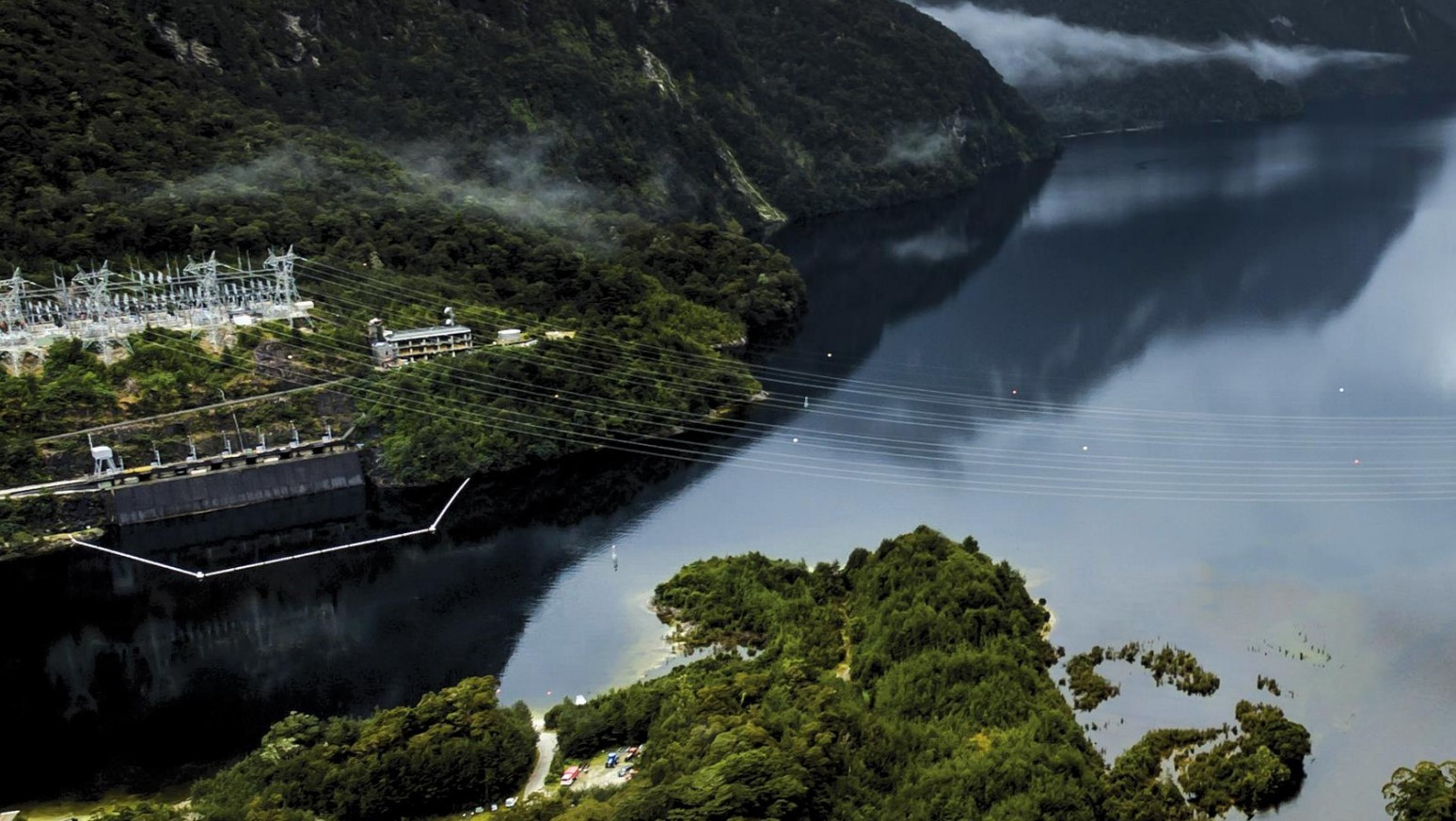 Hydroelectric dam with power lines over a lake surrounded by forested hills and mist.