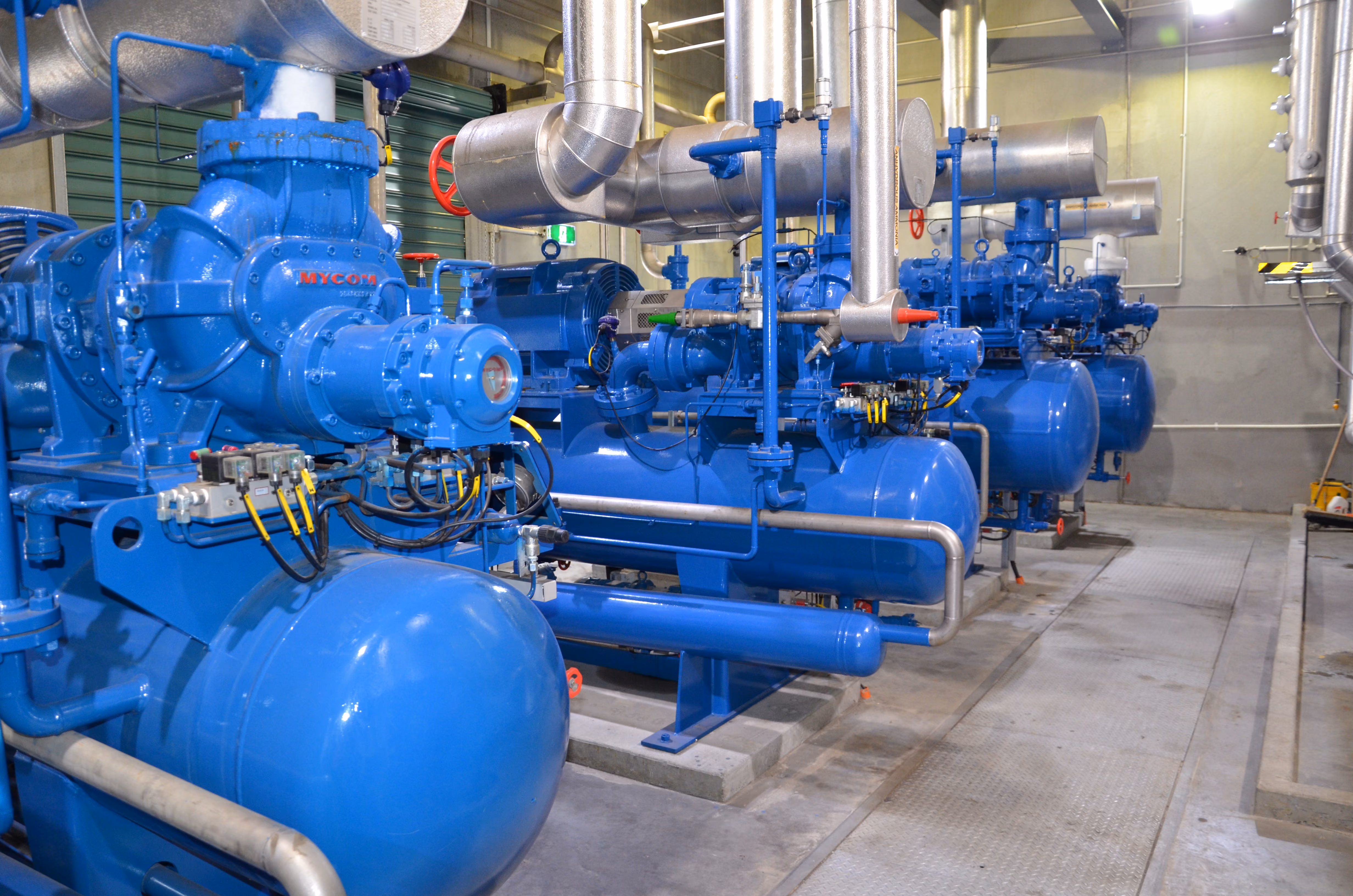 Row of large blue industrial compressors with connected silver pipes in a factory setting.