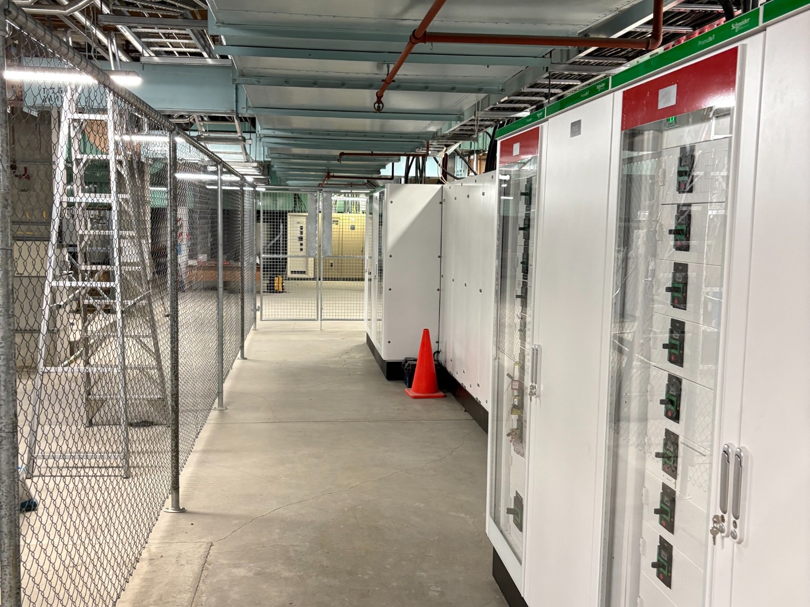 Industrial corridor with electrical control panels on the right and chain-link fencing on the left.