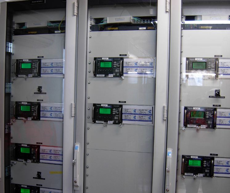 Electric control panel with multiple digital meters inside a grey metal enclosure with glass doors.