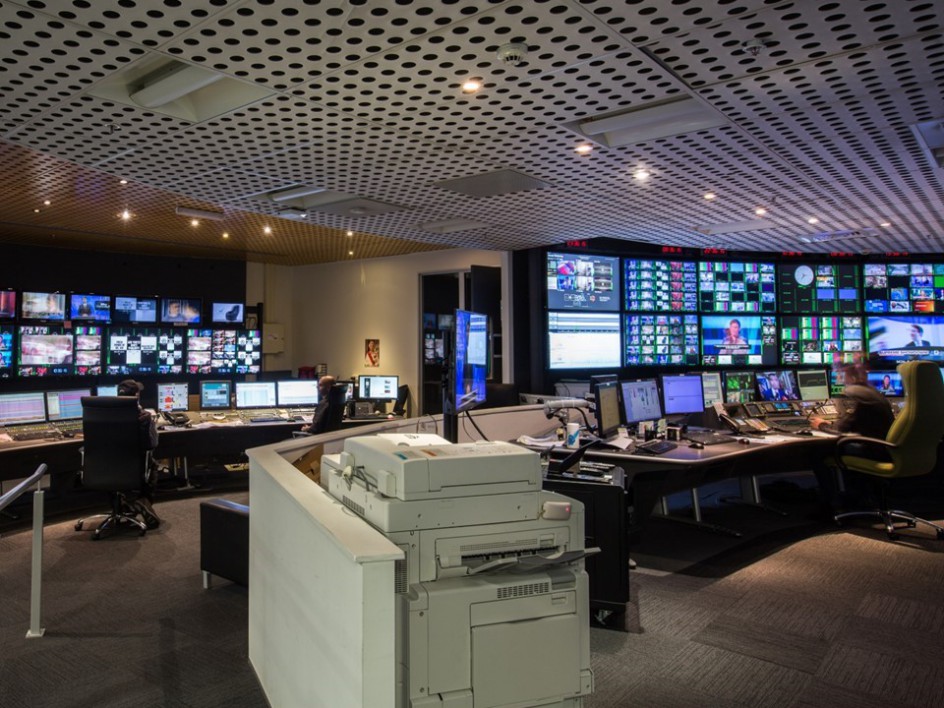 Broadcast control room with multiple monitors, control panels, and people working at desks.