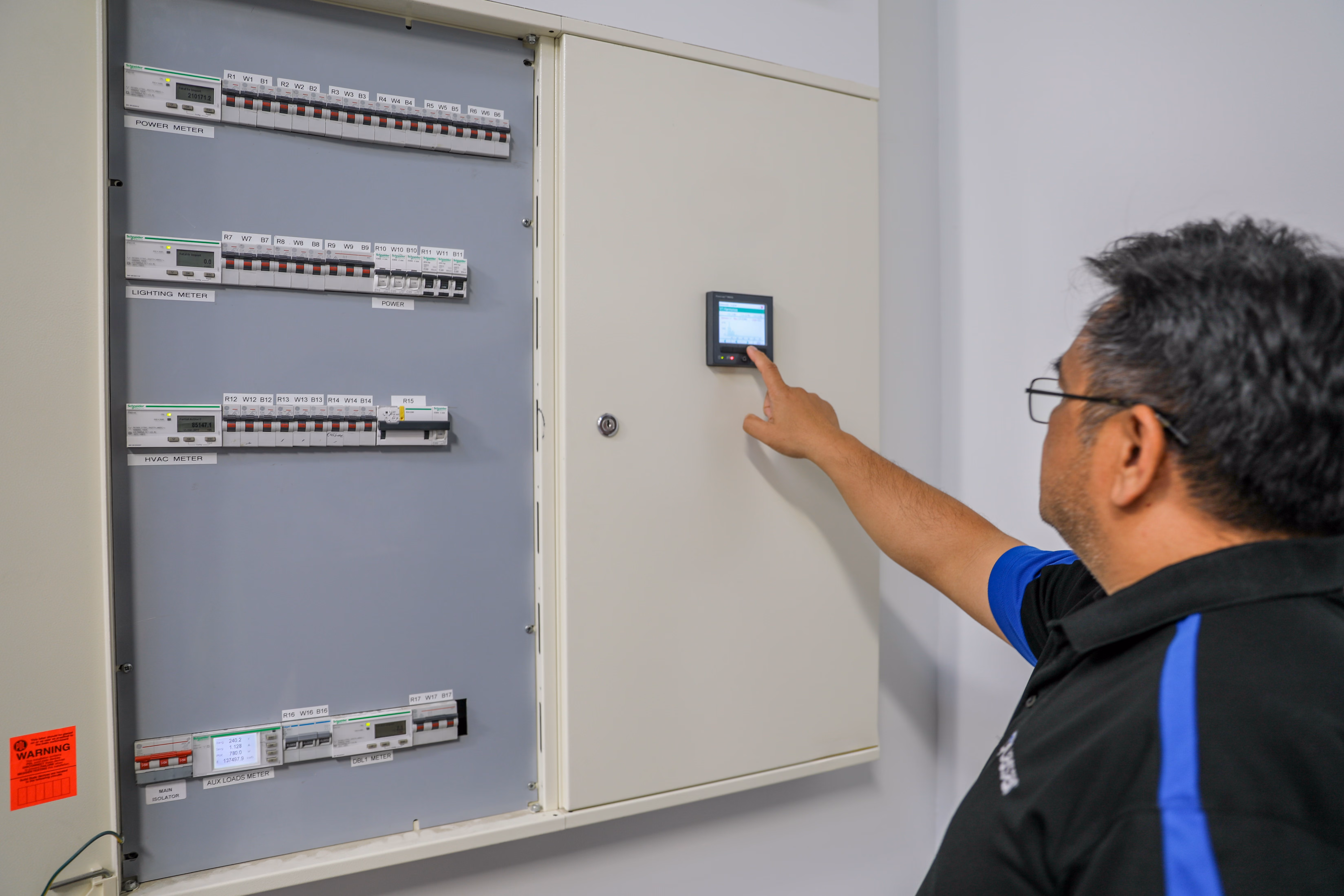 Man with glasses operating digital control panel on large electric meter and breaker panel.