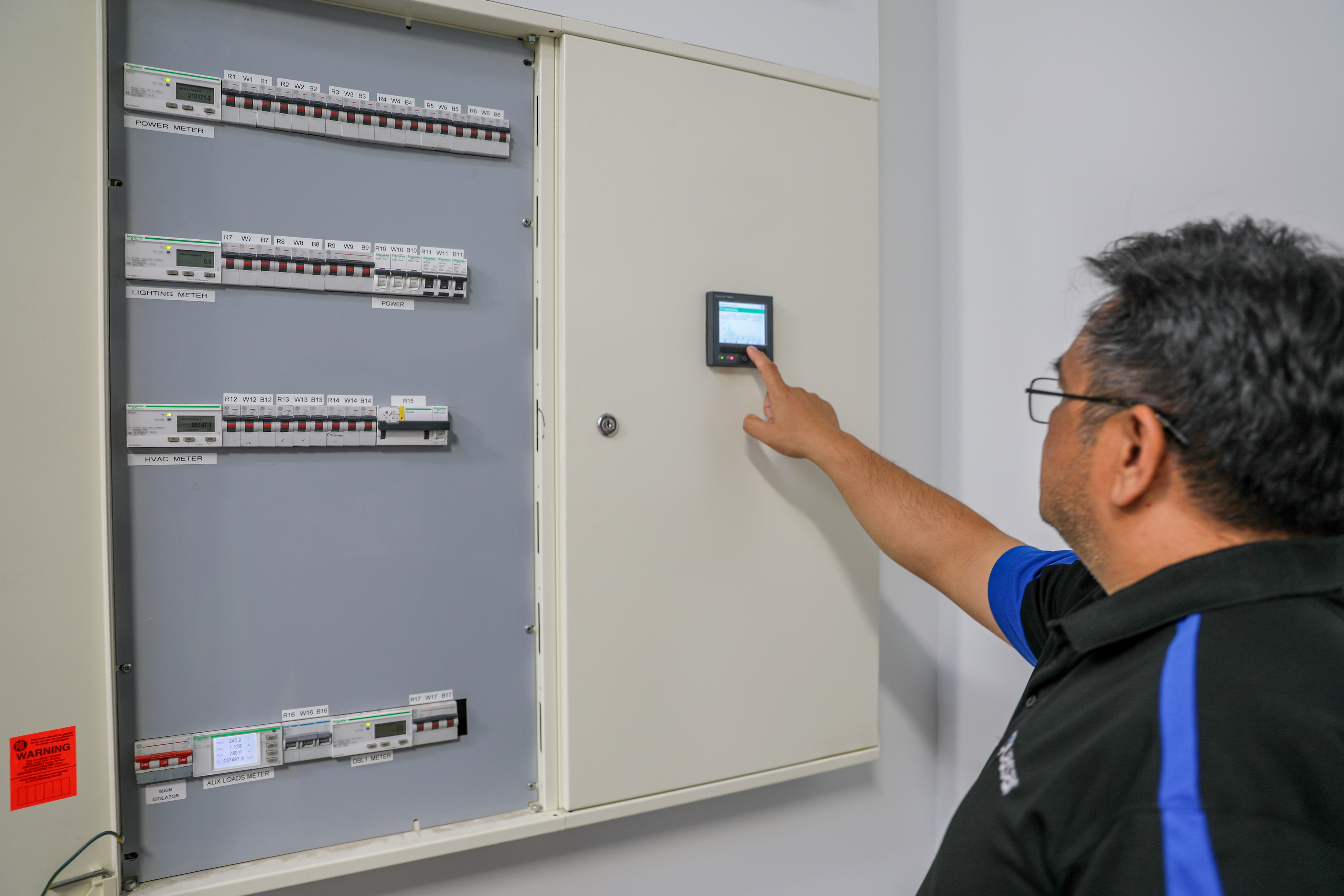 Man with glasses operating digital control panel on large electric meter and breaker panel.