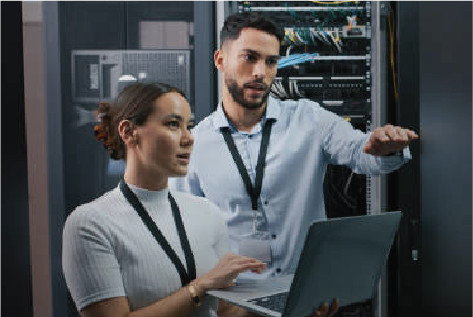 Two IT professionals discussing while one holds a laptop near a server rack in a data center.
