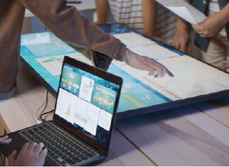 People interacting with a large touchscreen table while using a laptop with displayed charts and data.