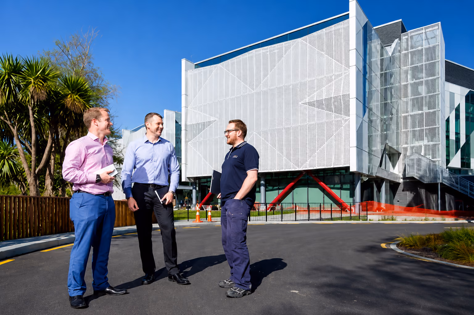 Three men standing outdoors near modern building, two in business attire, one in casual workwear.