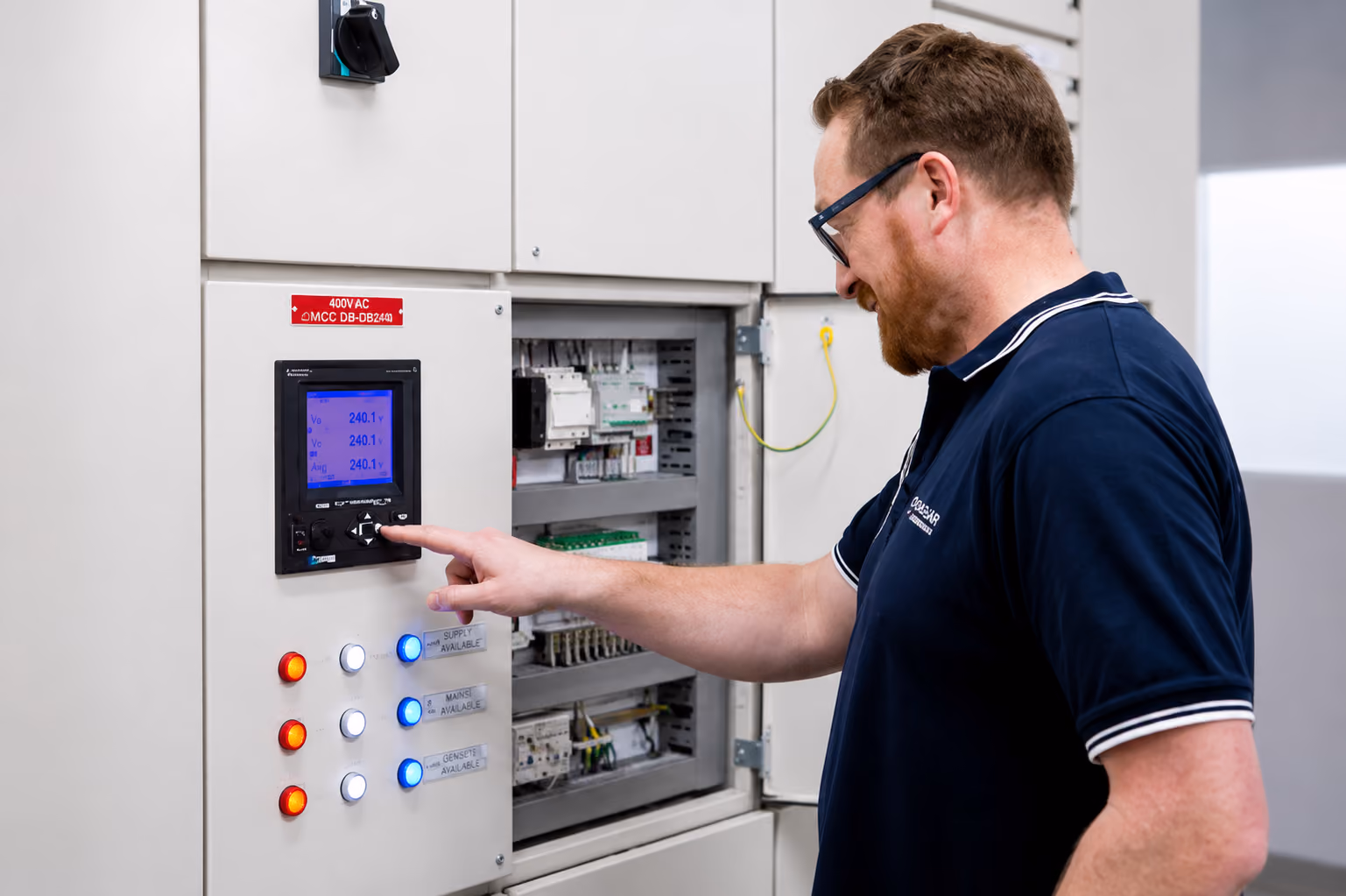 Man wearing glasses and navy shirt operates controls on an electrical panel with lit indicators.