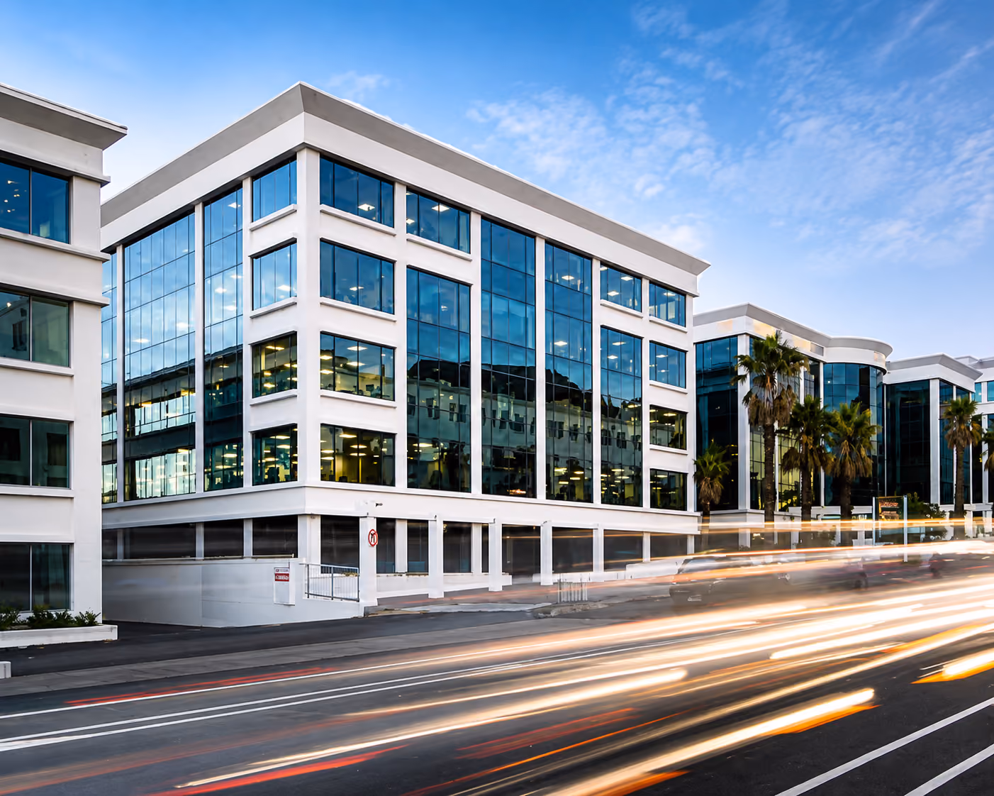 Modern office buildings with reflective glass windows and light trails of passing cars on street.