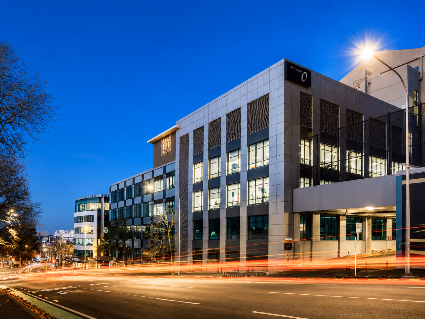 Modern office building at dusk with illuminated windows and light trails from passing cars.
